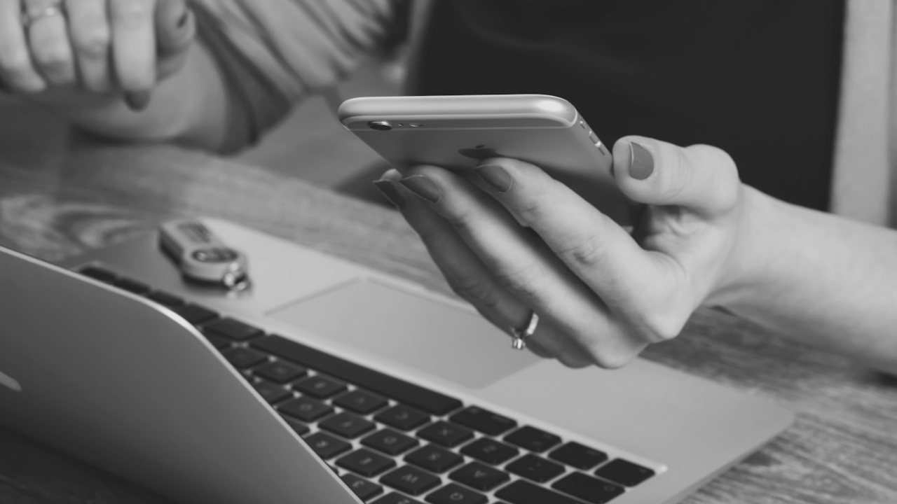 A person holding a smartphone above a laptop, with a USB drive nearby on the wooden table.