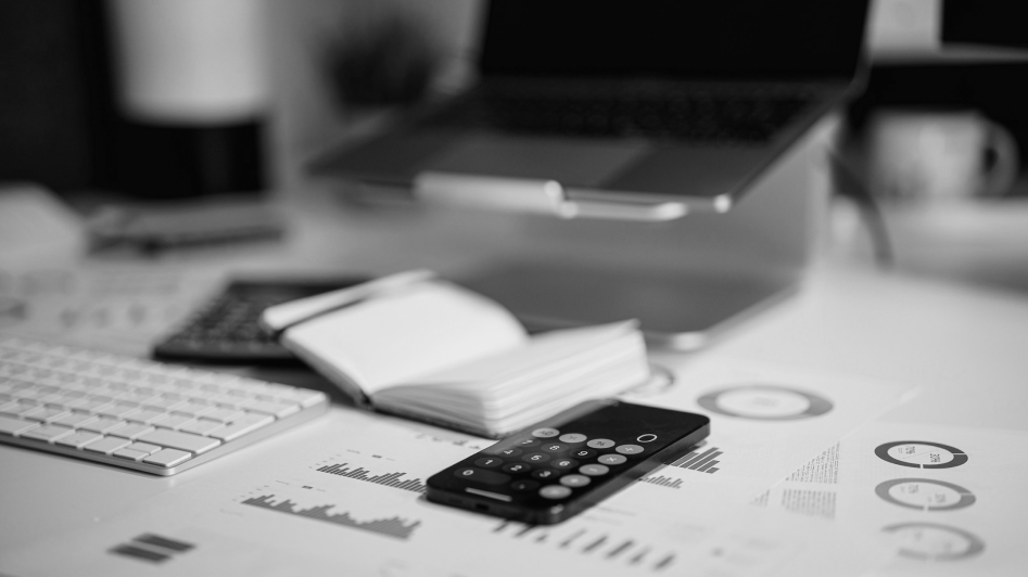 A workspace with a keyboard, smartphone showing a calculator app, notebook, and printed charts. Laptop in the background. Black and white image.