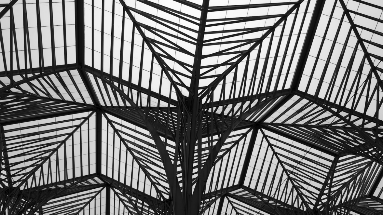 Architectural ceiling with geometric, lattice-like metal beams forming a symmetrical pattern against a glass backdrop.