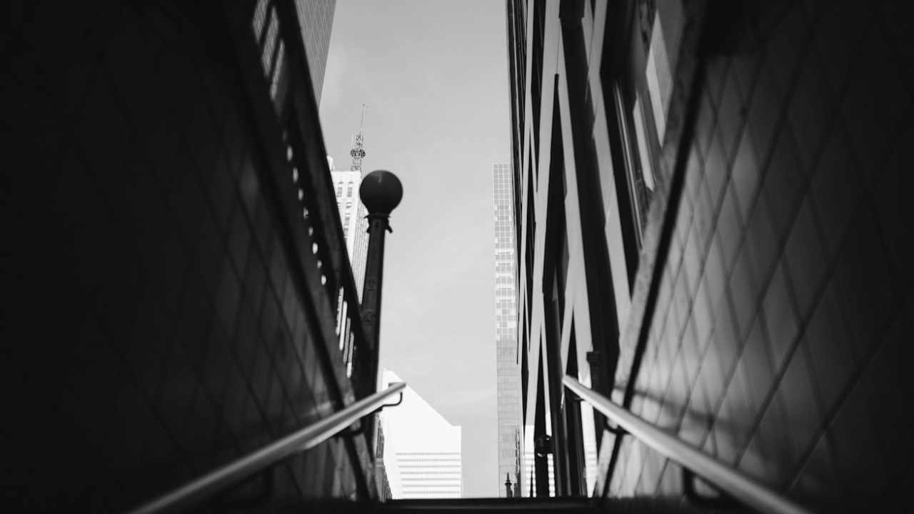 Black and white photo of a subway exit. Railings lead upwards to a cityscape with tall buildings in the background.