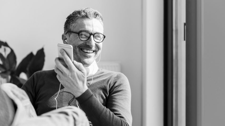 A man with glasses is smiling while holding a smartphone and wearing earphones, sitting by a window.