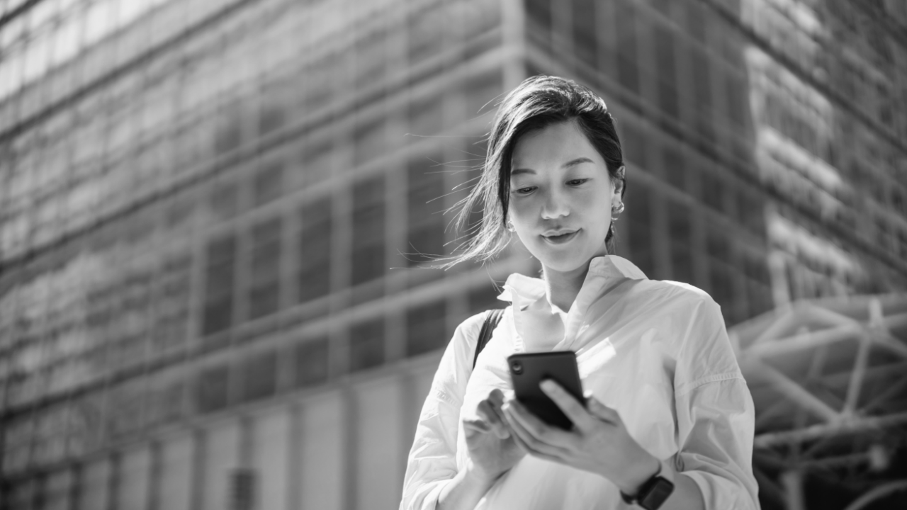 A woman in a white blouse is looking at her smartphone. She is standing outdoors in front of a large glass building. The image is in black and white.