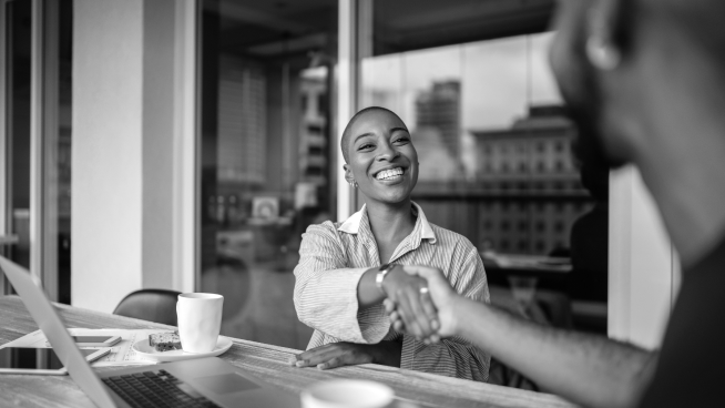 Two people shake hands across a table with laptops and coffee cups, smiling and sitting in an office setting.