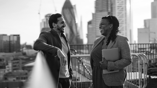 Two people talking and smiling on a rooftop with a city skyline in the background. One holds a laptop. Black and white photo.