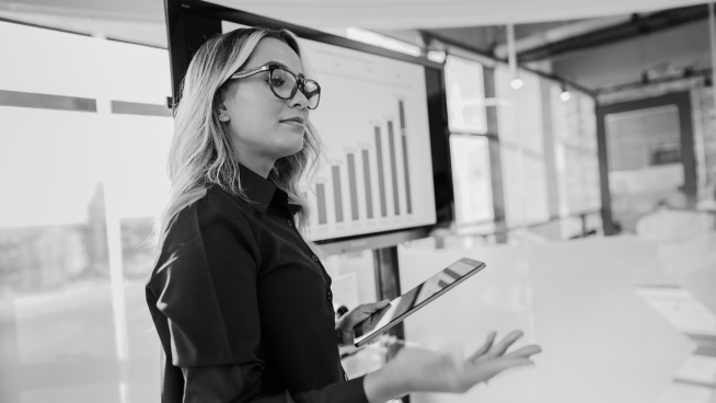 Woman with glasses holding a tablet, presenting in front of a screen displaying a bar chart.