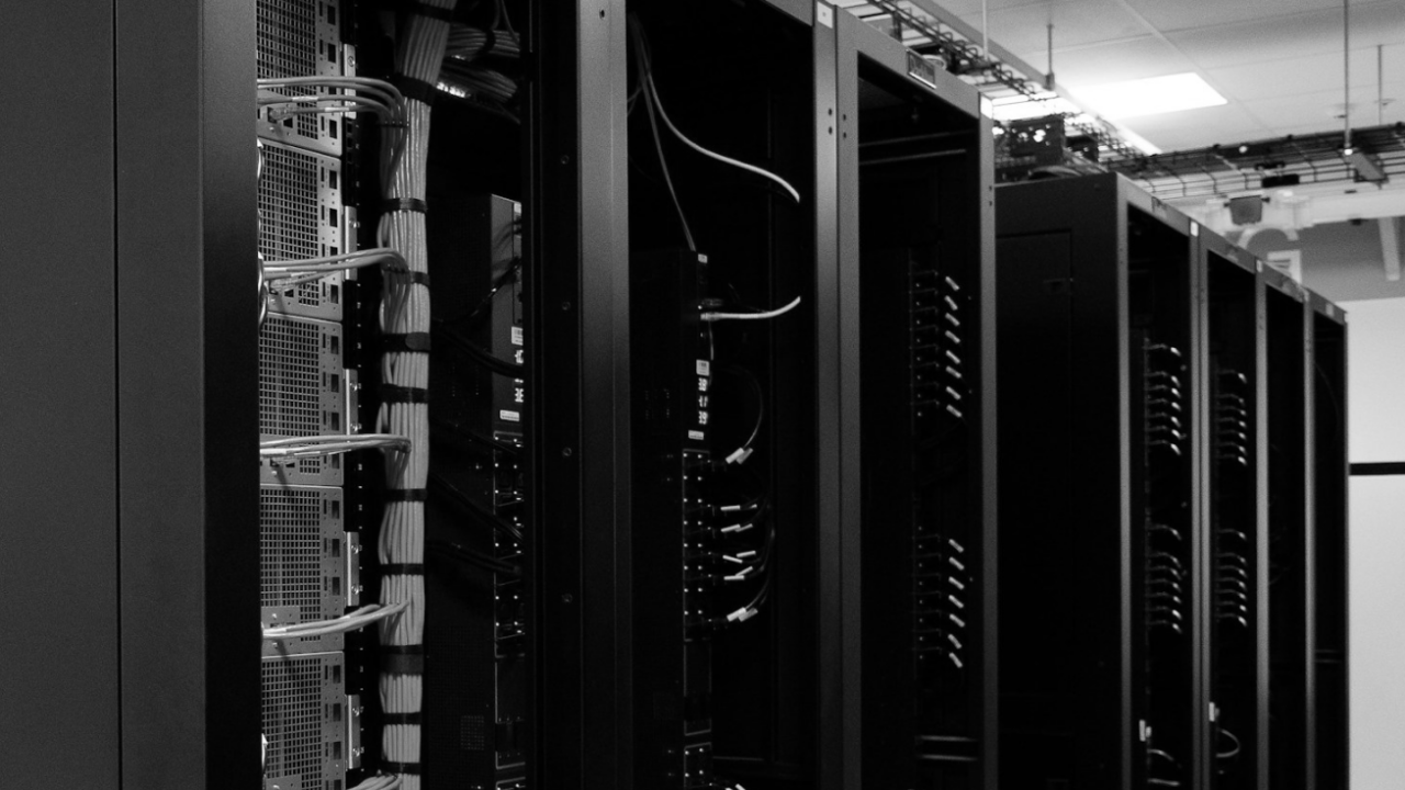 Black and white image of server racks in a data center with cables and equipment visible.