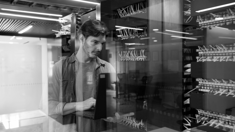 A person stands in a server room, using a laptop while examining network cables and equipment behind glass.