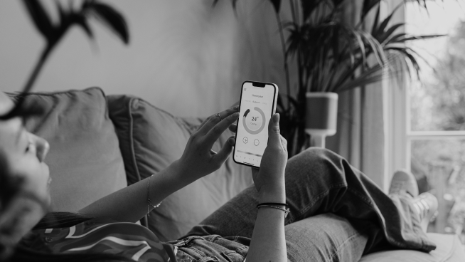 Person lying on a couch, adjusting settings on a smartphone with a temperature display. Plants and window in the background.