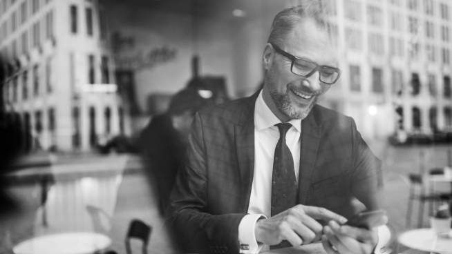 Man in a suit smiling and using a smartphone in a cafe, viewed through a window.