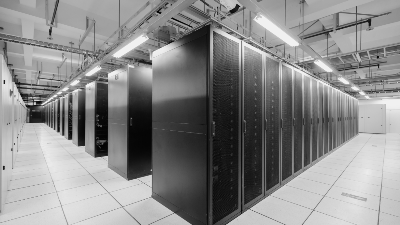 A black and white image of a modern data center with rows of server racks under bright overhead lighting.