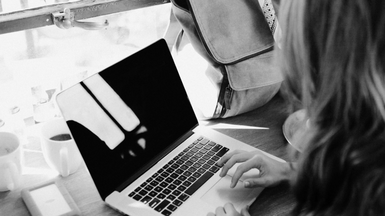 A person typing on a laptop at a wooden desk with two coffee cups and a backpack nearby.