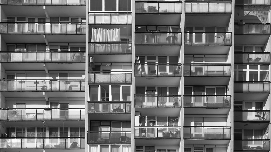 Black and white photo of a high-rise apartment building with multiple balconies, some featuring glass panels, plants, and laundry.