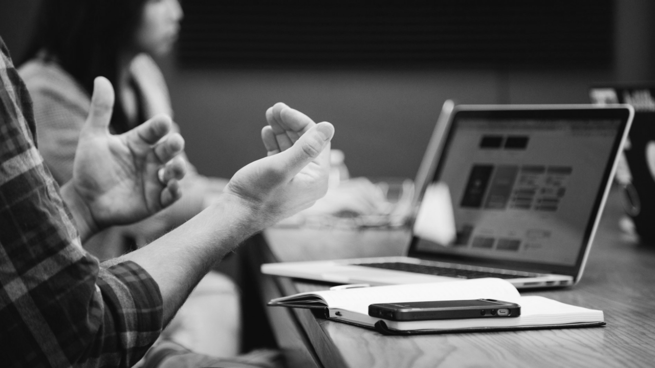 A person gestures with hands during a discussion at a table with a laptop, notebook, and phone. Another person is blurred in the background.