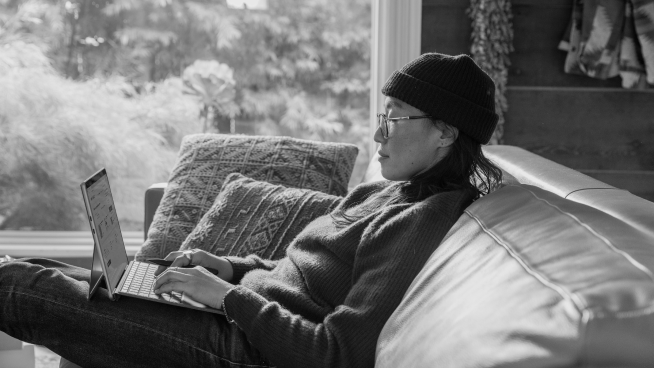 Person wearing a beanie and glasses, sitting on a sofa, typing on a laptop.