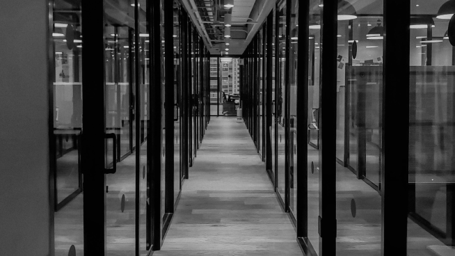 Black and white photo of a modern office hallway with glass walls and wooden floors, leading to a distant door.