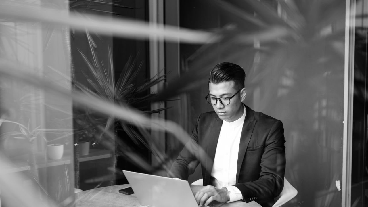 A person in a suit and glasses is seated at a desk, using a laptop, with plants partially obscuring the foreground.