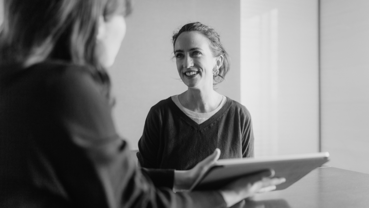 Two people are sitting at a table in conversation; one is holding a tablet. The image is in black and white.