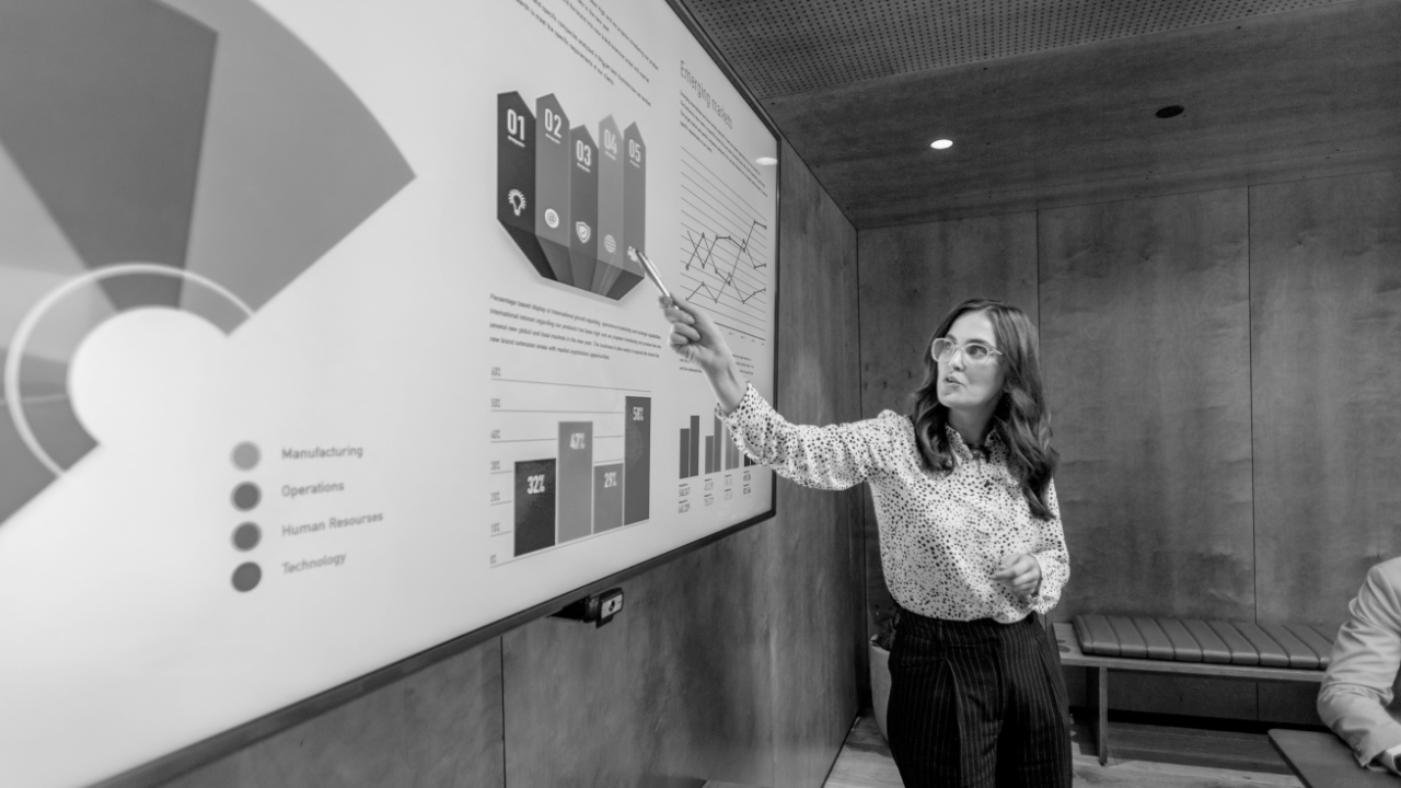 A woman stands in front of a screen, pointing at a colorful business chart during a presentation in a modern conference room.