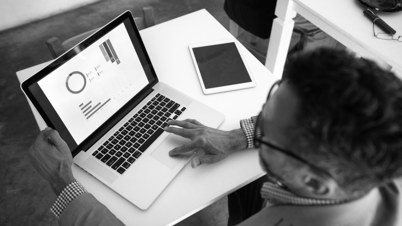 A person using a laptop displaying graphs, seated at a white table with a tablet nearby.